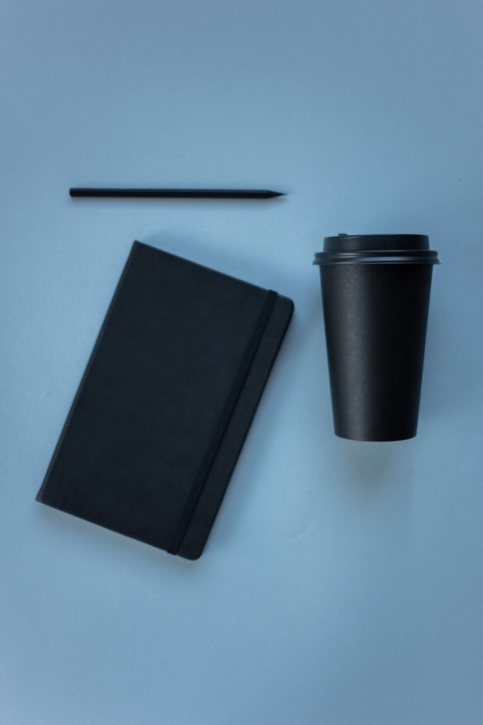 Simple flat lay of a black notebook, pen, and disposable coffee cup on a blue surface.