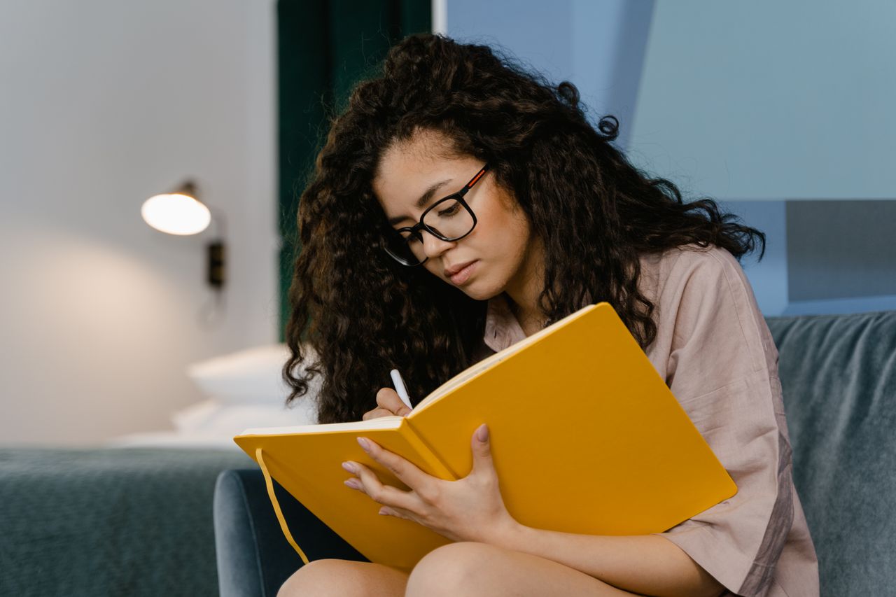 Young woman with curly hair writes in a yellow notebook, seated indoors.
