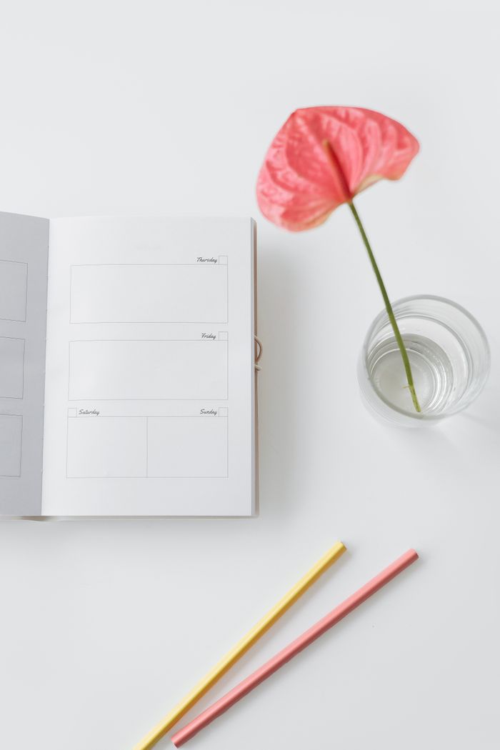 Flat lay of a minimalist workspace featuring an open notebook, a pink anthurium in a glass, and colored pencils.