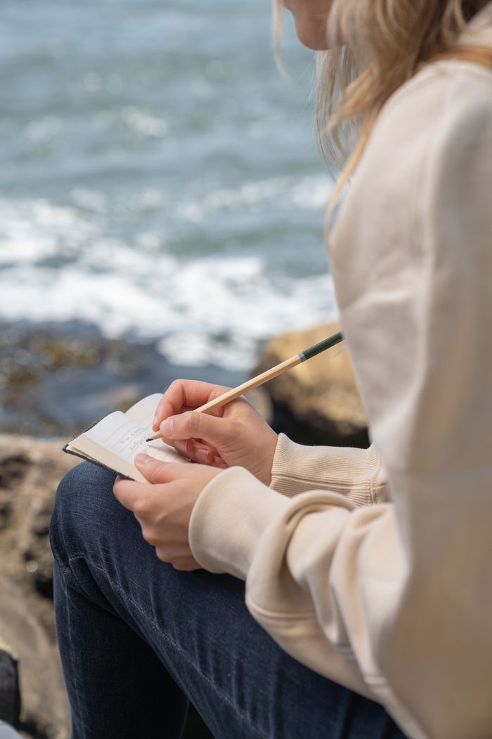 Close-up of a woman writing in a notebook while sitting by the ocean on a sunny day.